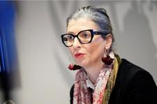 UN Special Rapporteur for the occupied Palestinian territories, Francesca Albanese speaks at a press conference. She is wearing thick, black rimmed glasses and silver hoop earrings with red cloth roses. She has on a black blazer with a red scarf.