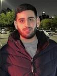 Sayfollah Musallet wearing a black puffer jacket over a gray t-shirt while standing in a parking lot at night and smiling at the camera
