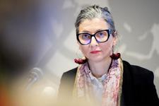 UN Special Rapporteur for the occupied Palestinian territories, Francesca Albanese speaks at a press conference. She is wearing thick, black rimmed glasses and silver hoop earrings with red cloth roses. She has on a black blazer with a red scarf.