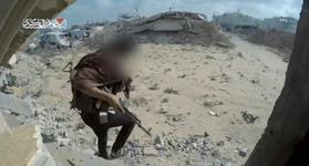 A Qassam fighter, whose face is blurred out, carries a rifle as they walk across dirt and rubble.