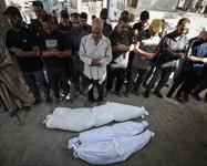 Men standing with their arms folded looking downward at three bodies wrapped in white linens on the ground