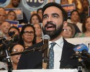 Zohran Mamdani speaking into several microphones at a lectern in front of supporters