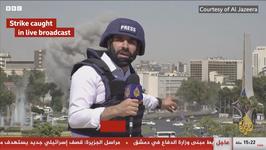 A reporter wearing a navy PRESS vest and helmet speaks to the camera as a missile hits a building behind him
