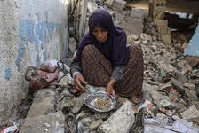 An elderly Palestinian woman with a long floral skirt and purple hijab squats as she picks out food scraps amid the rubble