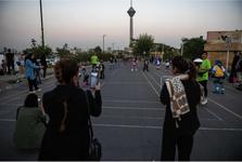 A woman is filming kids on her phone using their rollerblades on an open asphalt area in Tehran