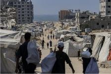Three people carrying white sacks face a dirt road that leads through many white tents and damaged buildings