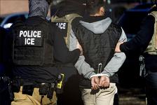 ICE officials wearing black vests escort a man wearing a black and gray hoodie with his hands bound behind his back in zip ties.