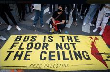 A person sits next to a large yellow banner on the ground that reads, "BDS is the floor, not the ceiling."