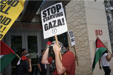 Demonstrators stand in front of a building holding Palestinian flags and carrying signs that read, "Stop Starving Gaza!"