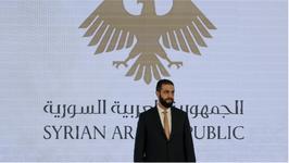 Syrian President Ahmed al-Sharaa wearing a dark suit and standing in front of a screen that reads, "Syrian Arab Republic"