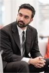 Zohran Mamdani wearing a black suit is seated in a chair leaning forward with his hands folded in front of him