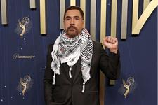Javier Bardem wearing a black suit in front of a blue and gold backdrop at the Emmy awards. He is wearing a keffiyeh and is posing with his fist raised.