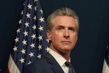 California Gov. Gavin Newsom wearing a navy suit, stands in front of an American flag
