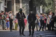 Two members of Hamas stand next to each other holding rifles while a group of children gather a short distance behind them
