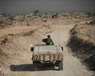 IDF solider rides in a military vehicle down a dirt road past destroyed buildings