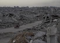 Two people wearing red sit on a cart pulled by a donkey as they ride down a road surrounded by the rubble of destroyed buildings