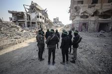 Members of Hama stand in a circle facing each other in a dirt road surrounded by destroyed buildings and rubble