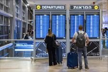 Travelers standing with their luggage looking at monitors displaying information for flight departures in Hebrew at an airport