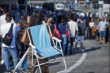 Thousands of people wait in line on a paved road. A woman holds a blue folding chair behind her.