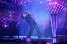 A person wearing all black sings into a microphone on stage with purple lights