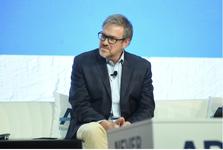 New York Times columnist Bret Stephens sits on a white couch wearing a dark blazer, light blue dress shirt, and khaki pants.