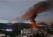 Orange flames and black smoke rise from hills located directly behind a cityscape