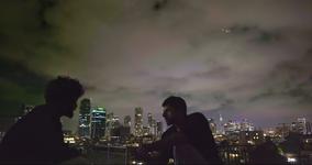 A scene from NAZA shows two men talking to each other in the dark with city lights shining from buildings behind them.