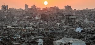 A few tents are seen among rubble and debris. Damaged buildings stand in the background.