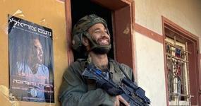 sraeli reservist and rapper Noam Tsuriely in military fatigues and helmet holding a rifle while smiling in the doorway of a building next to a poster of himself.