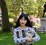 Author, RF Kuang, poses wearing a white blouse with a blue floral pattern while holding two books titled, "Babel" and "Katabasis"