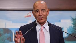 US ambassador to Turkey and special envoy for Syria Tom Barrack wearing a dark suit and pink necktie speaking at a lectern. Behind him is an image of a white building and the flag of Lebanon.