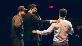 Three men stand on a stage. Mamdani (C) holds a microphone in his left hand and rests his right hand on the shoulder of Khalil (R).